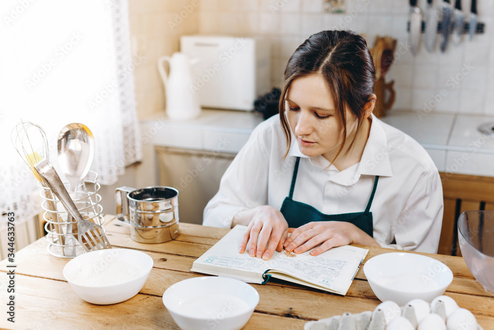 Housewife female chef in apron sitting at wooden table looking to the notebook with recipes in a kitchen. Cooking at home concept. Housewife female chef in apron sitting at wooden table looking to the notebook with recipes in a kitchen. Cooking at home concept.