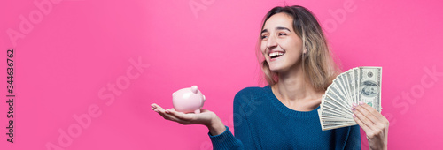 Girl puts in the piggy bank one hundred dollar bills. Young girl over pink background holding piggy bank and rejoices gesturing. The concept of reliability of cash investments and insurance.