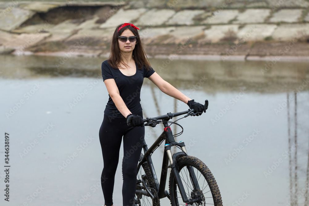 Fototapeta premium Girl on a mountain bike on offroad, beautiful portrait of a cyclist in rainy weather, Fitness girl rides a modern carbon fiber mountain bike in sportswear. Close-up portrait of a girl in red bandana.