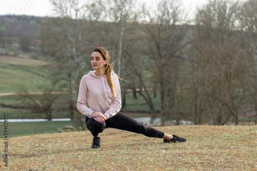 A young slim athletic girl in sportswear performs a set of exercises. Fitness and healthy lifestyle against the background of green spring pasture hills.