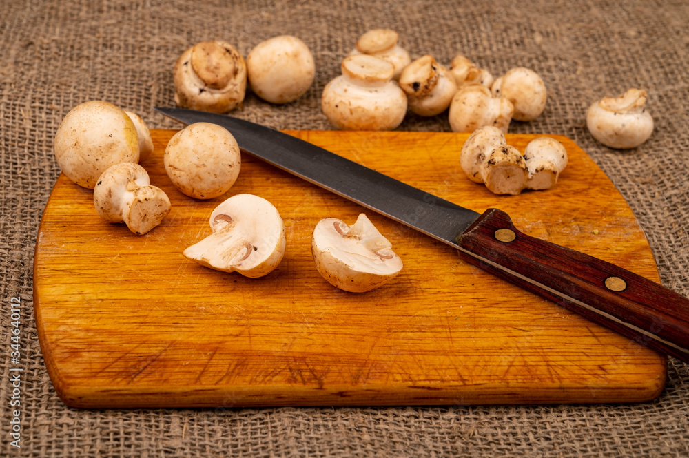 Halves of cut young mushrooms and a knife with a wooden handle on a wooden cutting Board and mushrooms scattered on a background of coarse homespun fabric. Close up. Halves of cut young mushrooms and a knife with a wooden handle on a wooden cutting Board and mushrooms scattered on a background of coarse homespun fabric. Close up.