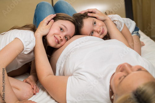 children, little girls listen to the belly of a pregnant mother lying on her back at home on the bed