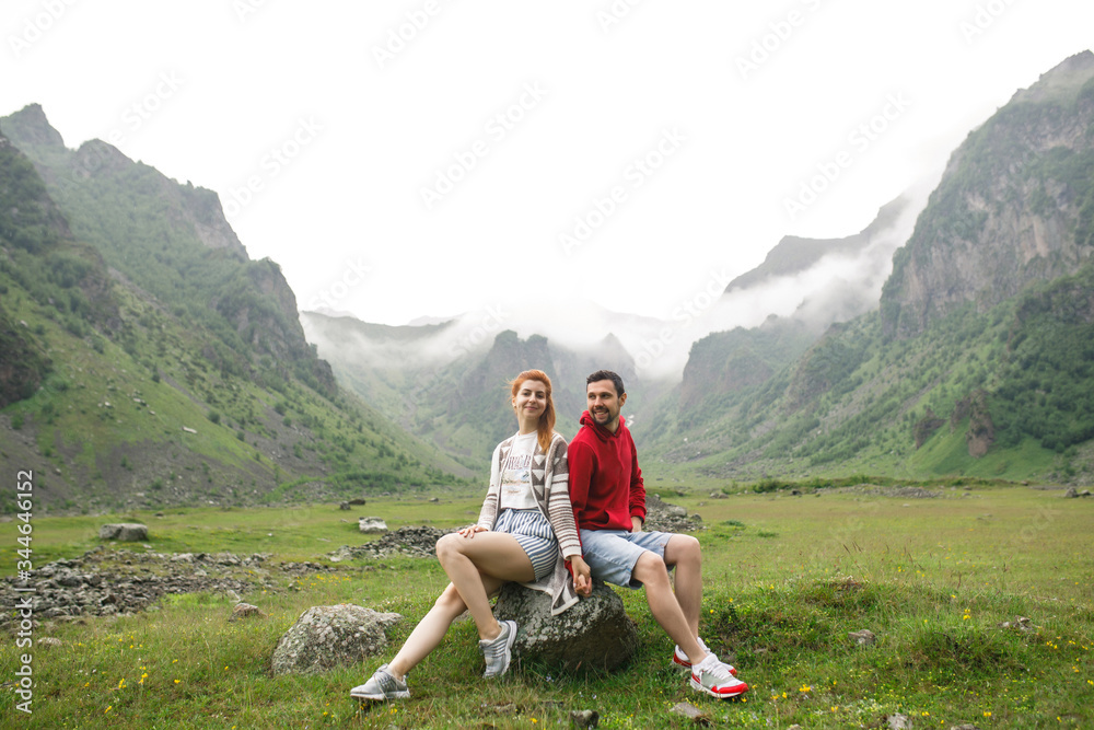 Naklejka premium young beautiful couple of travelers girl blonde in a white T-shirt, a man in a red sweater sitting on a background of mountains in Georgia