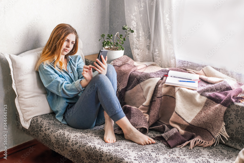 Obraz premium Pretty,caucacian,young girl resting on a sofa holding,using cellphone.Work by mobile,messaging,online shopping,distance communication.Isolated indoor while quarantine time.Stay at home concept.