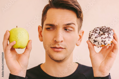 Portrait of funny uncertain fitness man hold fresh apple and sweet donut snack in hands. Young sports man struggling between healthy fruit and unhealthy junk food. Motivation for dieting, weightloss