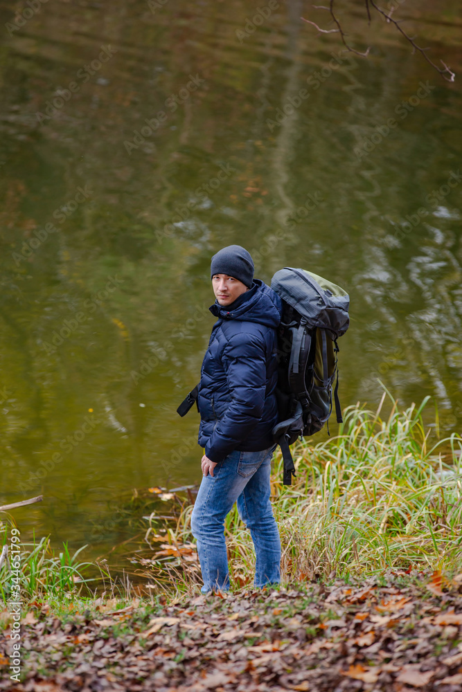 Tourist in a jacket and jeans looks at the camera near the river. Autumn landscape background.