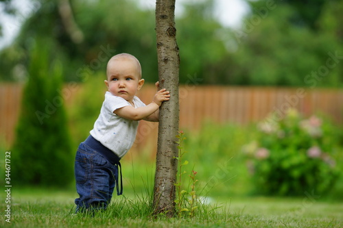 Side portrait of little funny cute baby in jeans and white shirt holding on tree trunk, bright green background