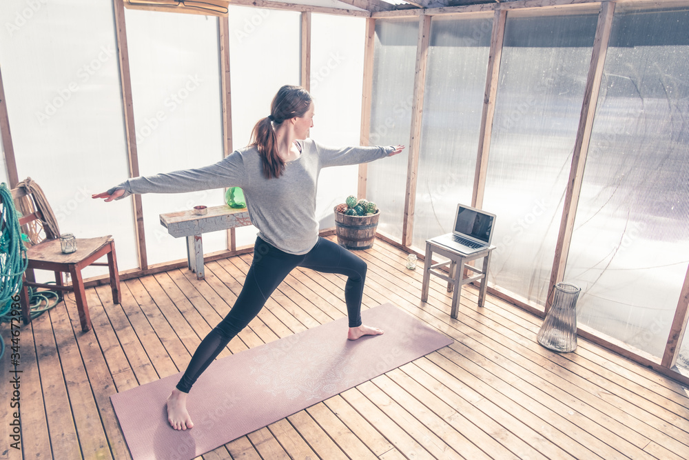 Fototapeta premium Young woman is doing Warrior pose during yoga practice at her home studio due to COVID-19 restrictions. She follows online yoga course on the computer. Healthy lifestyle photo and urban interior. 
