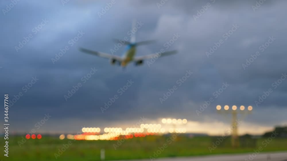 Defocused view of landing passenger plane, few cars pass by on road. Low angle shot, plane silhouette fly by against cloudy sky. Round lights of approaching lighting system at end of runway Defocused view of landing passenger plane, few cars pass by on road. Low angle shot, plane silhouette fly by against cloudy sky. Round lights of approaching lighting system at end of runway