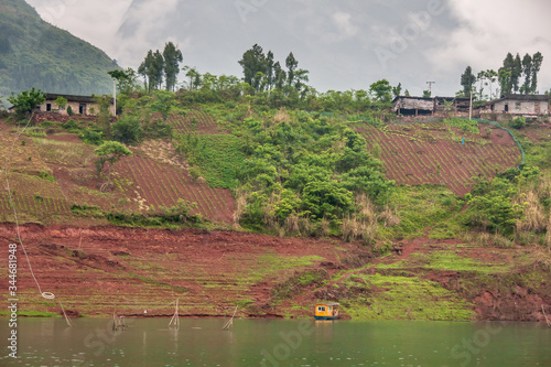 Wuchan, China - May 7, 2010: Dicui or Emerald Gorge on Daning River. Brown farmland with green vegetation on slope descending to green river with fish pen, and cabin boat under cloudscape. photo