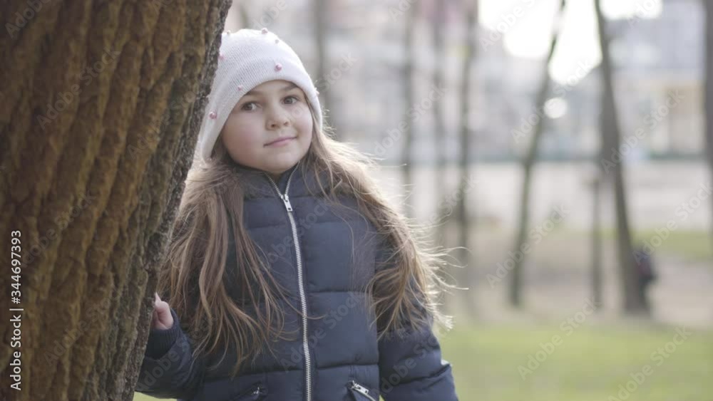Portrait of surprised Caucasian little girl looking at camera and hiding behind the tree trunk. Joyful kid having fun outdoors on sunny spring day. Nature, lifestyle, relaxation.