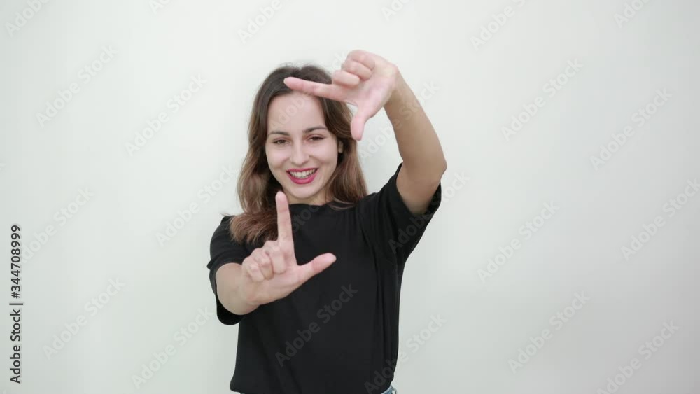 Cute Young Brunette Woman In Black T-Shirt, Blue Jeans With Belt On Gray Background, Happy Girl Smiling And Hands Imitating A Photo Frame, Make Virtual Photo With Fingers