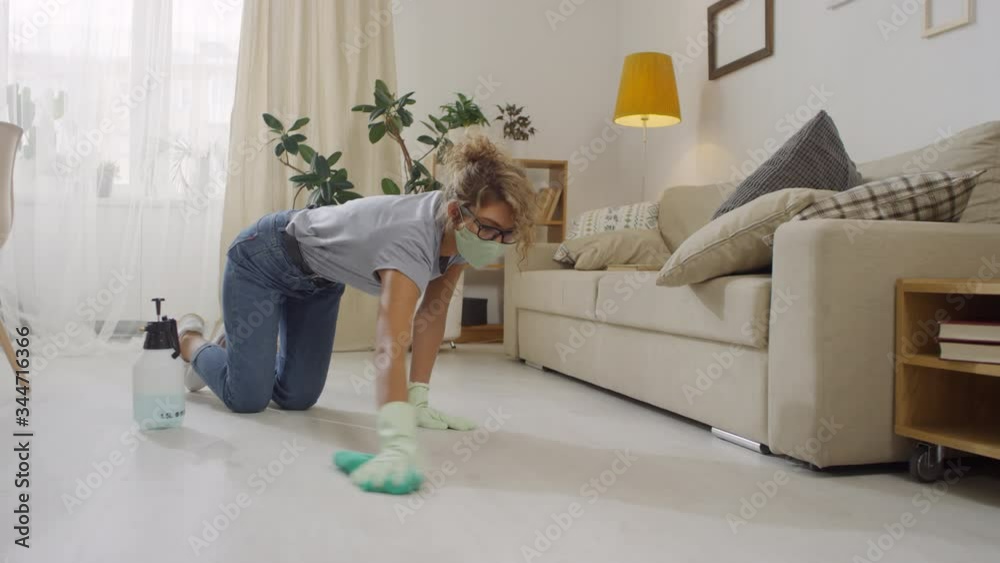 Medium shot of young woman in glasses, face mask and gloves kneeling and scrubbing floor with sanitizing liquid at home Medium shot of young woman in glasses, face mask and gloves kneeling and scrubbing floor with sanitizing liquid at home