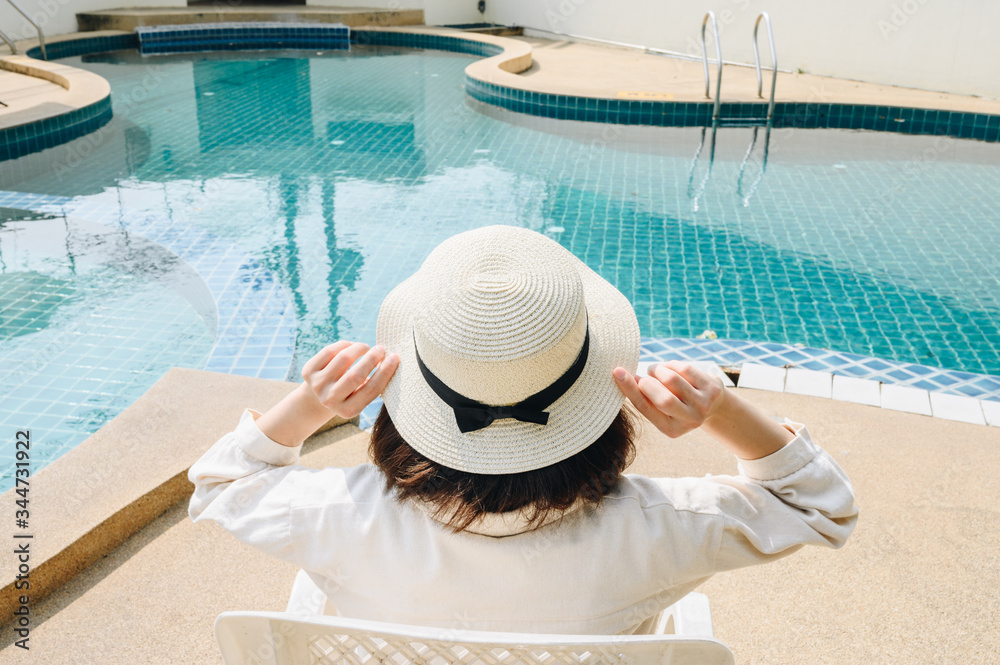 Obraz premium Back view of woman relaxing and sitting nearly swimming pool in hotel. This pool designed to hold water for leisure activities. Conceptual of tourist woman lifestyle.