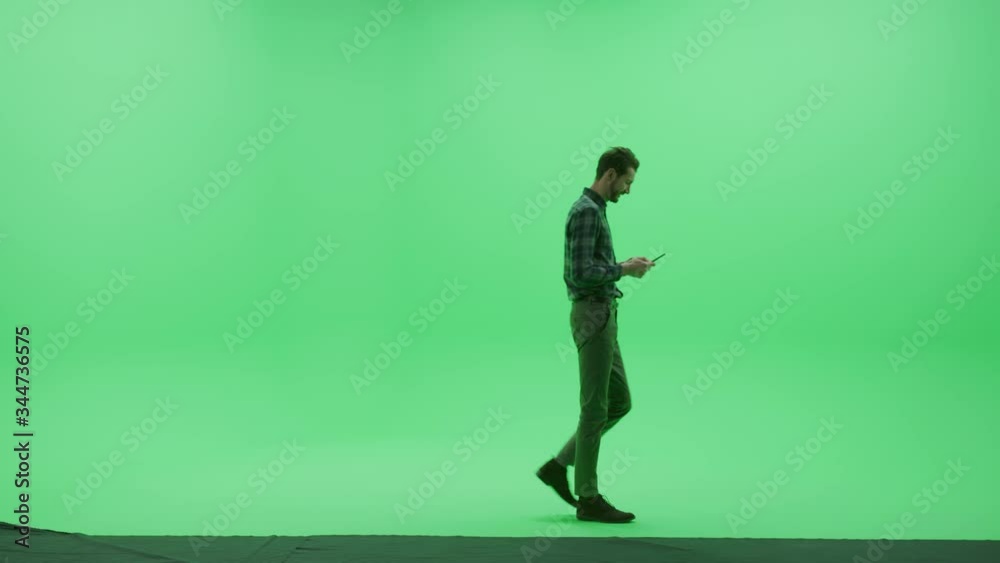 Green Screen Chroma Key Studio: Strong Man Wearing Casual Clothes, Uses Smartphone Device, Writes Email, Does Remote Online Work, Browses Through Internet Walks Across Room. Side View Camera Shot