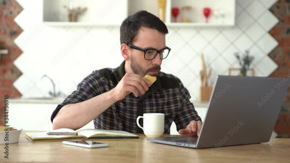 Serious young man working on the laptop while eating a snack. Working from home at the kitchen