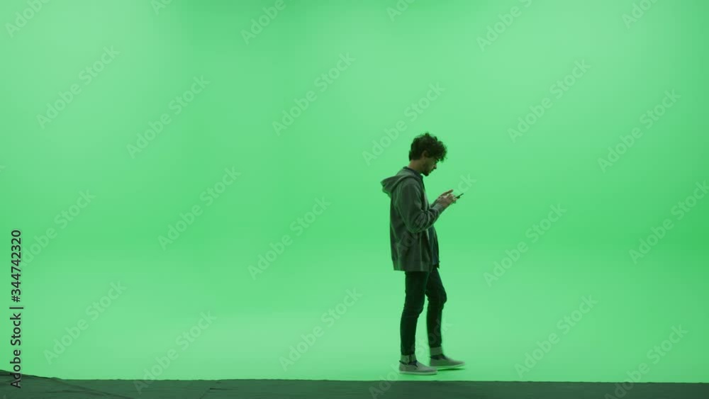Green Screen Chroma Key Studio: Handsome Young Man Wearing Casual Clothes, Uses Smartphone, Does Remote Online Work, Browses Through Internet and Social Media Walks Across Room. Side View Camera Shot Green Screen Chroma Key Studio: Handsome Young Man Wearing Casual Clothes, Uses Smartphone, Does Remote Online Work, Browses Through Internet and Social Media Walks Across Room. Side View Camera Shot