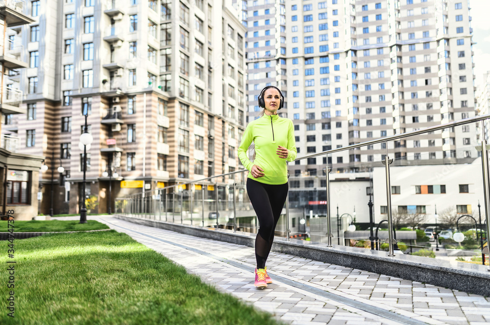 Morning jog. Young woman in sportswear runs among residential skyscrapers, she is listening music in headphones