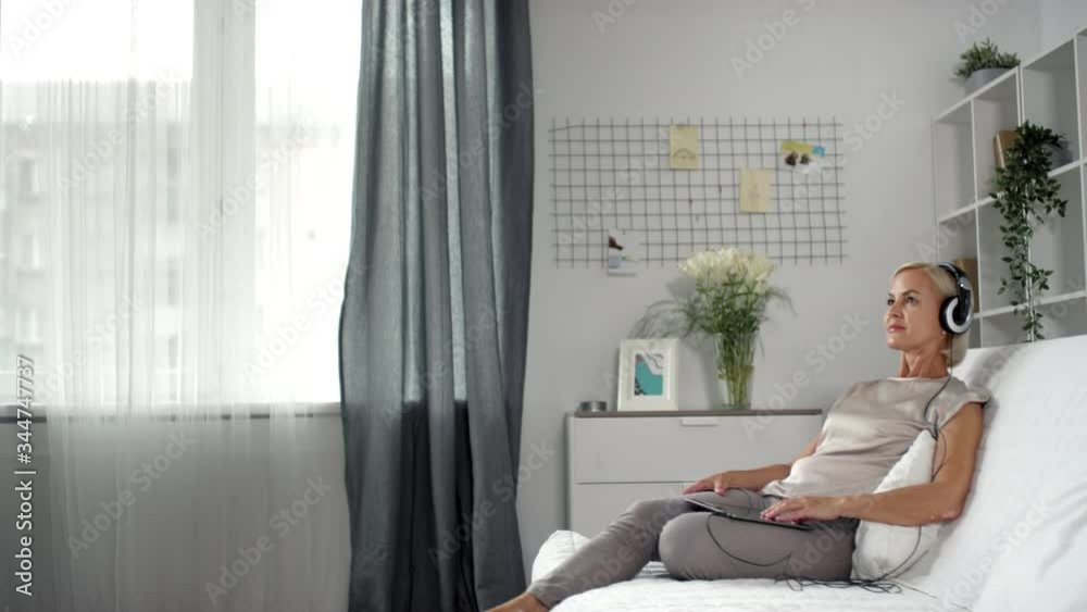 Beautiful mature woman sitting on couch in the living room and listening to good music with headphones on digital tablet Beautiful mature woman sitting on couch in the living room and listening to good music with headphones on digital tablet
