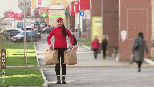 Courier with a packages of food on the street.
A girl from a food delivery service walks up to two bags of groceries and picks them up.
From the packages stick out: baguettes, milk, water, bananas.
