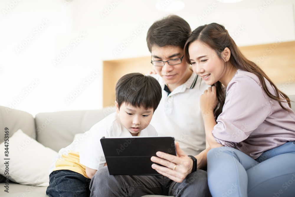 Asian family happy using tablet computer together for watch social media and sitting on gray color sofa in living room with copy space, parent advice kid for internet online concept. Asian family happy using tablet computer together for watch social media and sitting on gray color sofa in living room with copy space, parent advice kid for internet online concept.