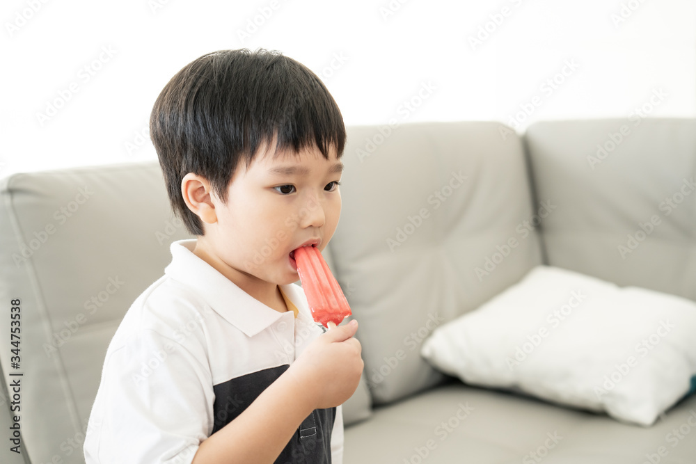 Asian cute little boy standing eating red color ice-cream strawberry or watermelon stick in living room with copy space