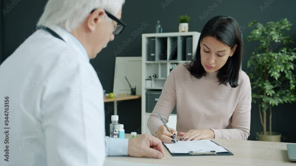 Female patient is talking to male doctor then signing medical chart during visit to hospital, cheerful people are discussing treatment and diagnosis. Female patient is talking to male doctor then signing medical chart during visit to hospital, cheerful people are discussing treatment and diagnosis.