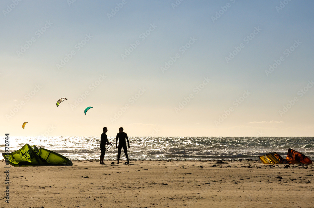 Silhouettes of two kitesurfer guys standing on the ocean beach with waves and multi-colored sails.