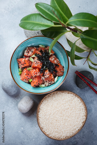 Poke bowl with salmon and a coconut shell with raw rice, above view on a light-blue stone background with green ficus leaves and pebbles