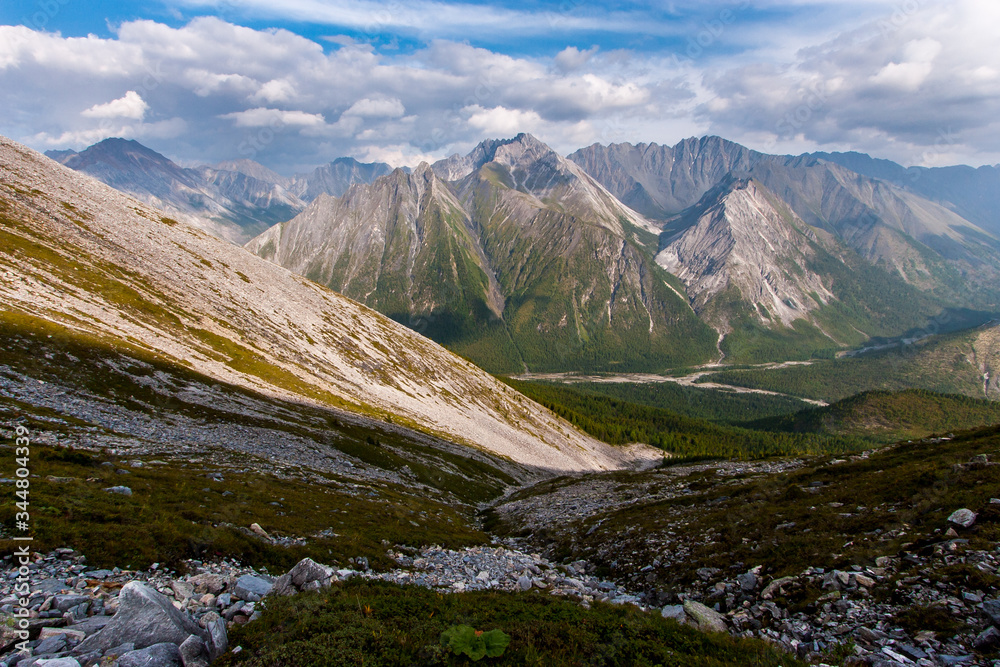 View from the mountainside into a valley with a green forest, a river and mountain ranges behind it. Rocky scree on a slope. Clouds on the blue sky. Horizontal.