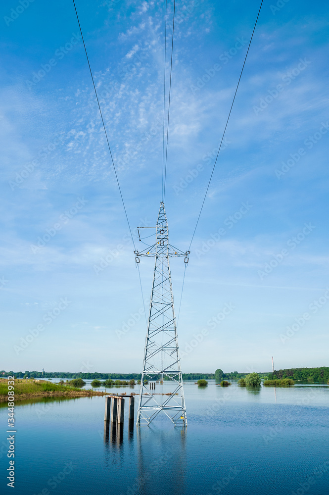 The high voltage power lines on water. Blue sky and green trees during river flood via pylon tower.