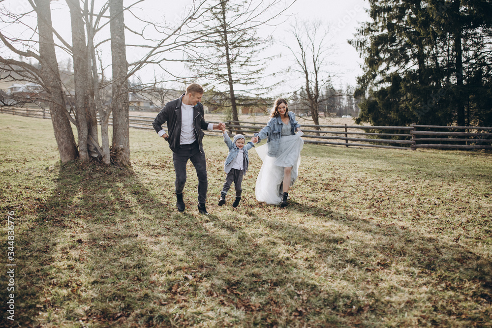 Fototapeta premium Stylish family in the autumn forest. A guy in a leather jacket and a young girl in a gray-blue dress play with his son in a meadow of a pine forest in the mountains against the background of a fence 