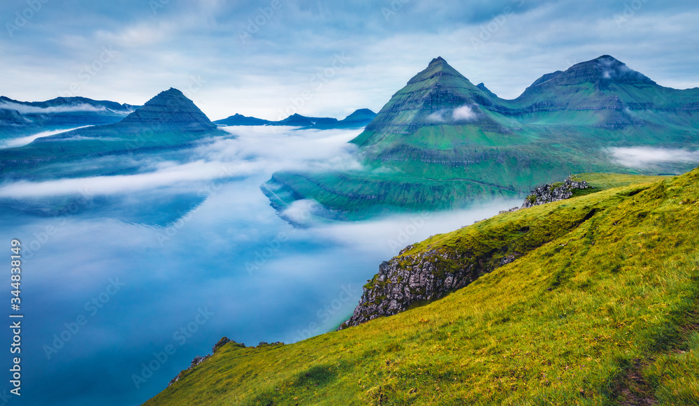 Dramatic morning view of Faroe Islands with low clouds, Denmark, Europe with low clouds. Adorable summer scene of Eysturoy island, Funningur village location. Beauty of nature concept background.