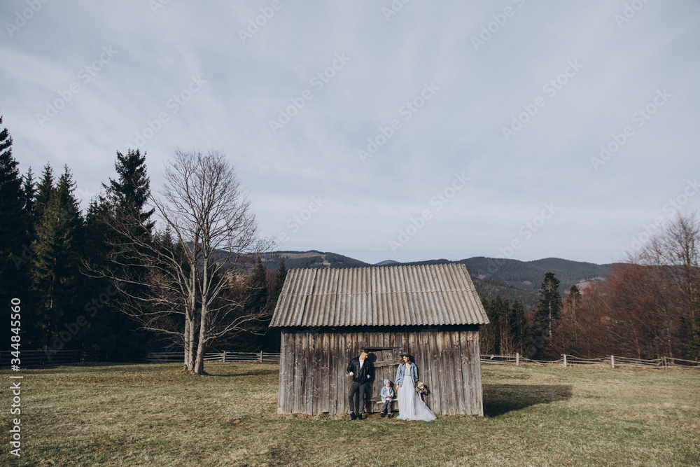 Fototapeta premium Stylish family in the autumn mountains. Stock photo. A guy in a leather jacket and a young girl in a gray-blue dress with his son on the background of an old wooden barn at sunset