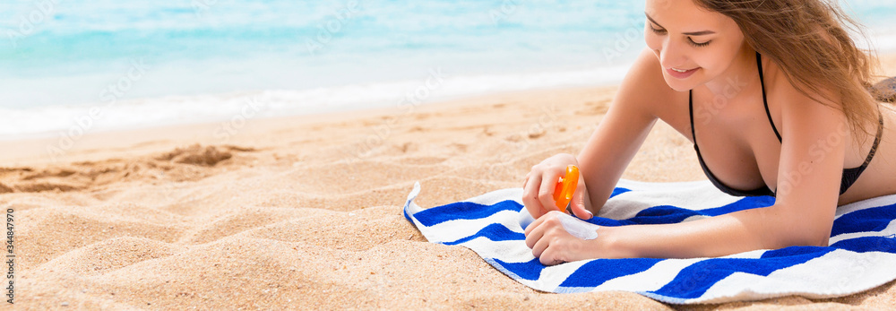 © sosiukin - Young happy woman is resting on the towel near the sea and protects her skin on the hand with sunscreen from the spray