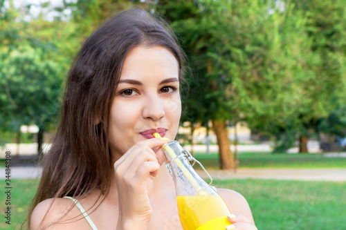 Attractive girl is drinking fresh juice through the straw outdoors. Pretty woman is holding a bottle of cold lemonade