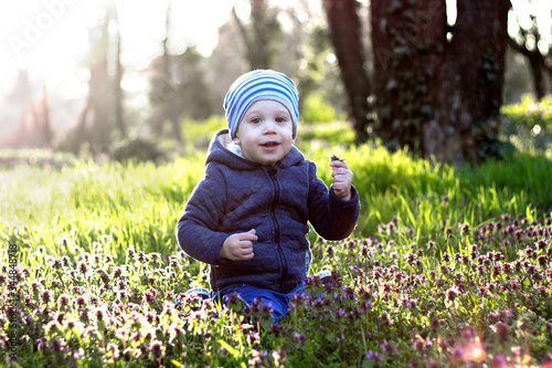 Happy little baby, kid smiling, sitting, holding, collecting flower in forest.