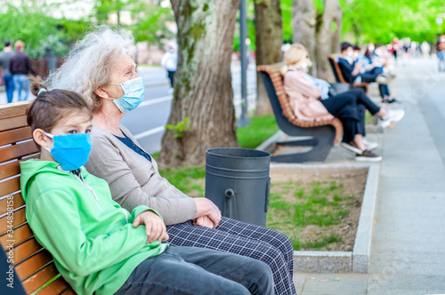Grandmother with her grandson in respiratory protective masks in an crowded promenade town sitting on a bench. Coronavirus covid-19 quarantine attenuation. Health people.Outdoors walk
