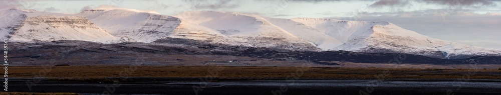 A panorama and wide angle shot of snow capped rocky mountains located on the edge of Hvalfjordur, Iceland. Wispy clouds cover the peaks as the sun rises lighting the mountainside. 