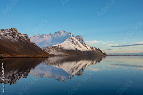 A snow capped mountain perfectly reflected in still blue water.
