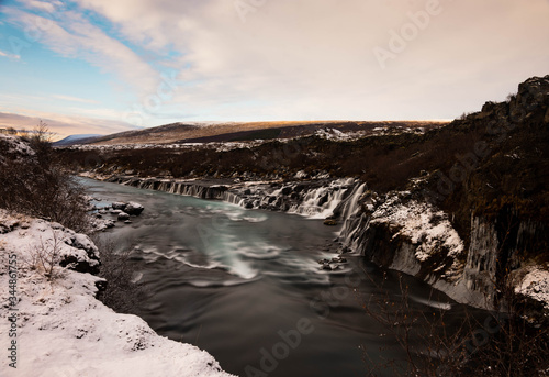 A turquoise blue river flows through a snowy autumnal landscape with water cascading over lava rock formations.