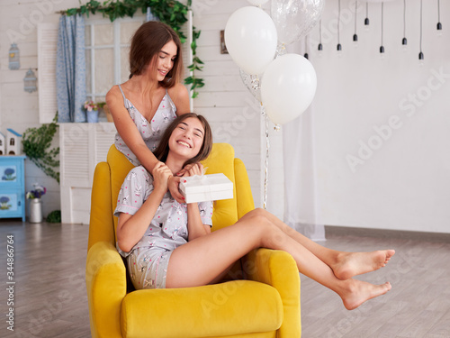 Two young brunette girls, wearing light grey cotton pajamas, sitting on yellow armchair in light apartment. Birthday celebration. Pretty woman is congratulating her sister, giving her wrapped present.