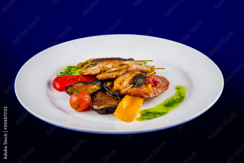 Turkey medallions stuffed with herbs on skewers with grilled vegetables and pesto sauce on a white plate on a blue background.