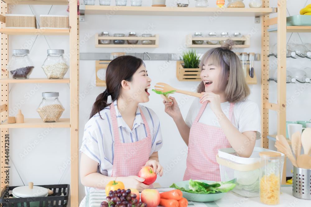 Two beautiful Asian women, Thai people wear casual clothes and wear pink aprons, helping each other cook and have fun tasting food in the kitchen together.