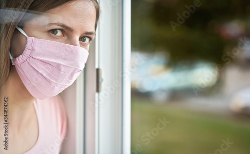 Young woman in pink cotton face virus mask standing behind window glass pane, touching it with hand, looking sad. Quarantine or stay at home to be safe during coronavirus covid-19 outbreak concept