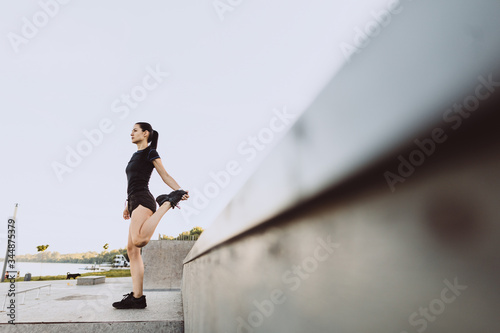 Athletic girl dressed in black, warms up before running, on city beach in the morning