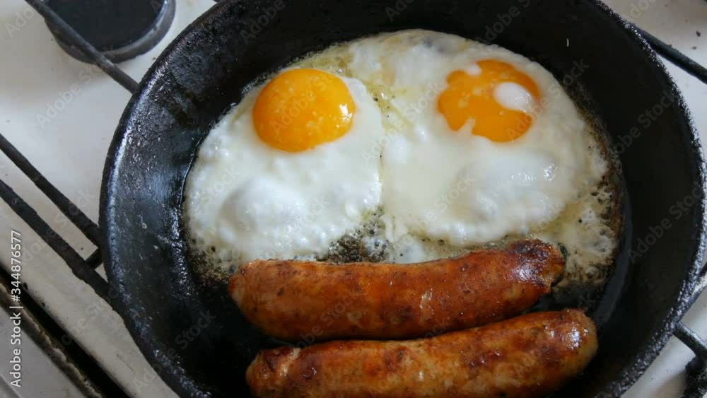 Funny breakfast in the pan in the form of a face and a smile with eyes. Two fried eggs and thick Bavarian sausages fried in a vintage cast iron skillet on a gas stove. Funny breakfast in the pan in the form of a face and a smile with eyes. Two fried eggs and thick Bavarian sausages fried in a vintage cast iron skillet on a gas stove.