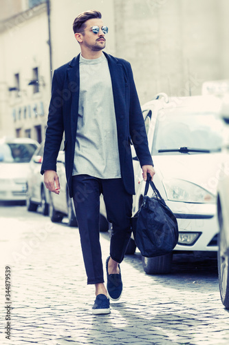 Fashionable young man walking in the street in the city. Near to white taxi cars. With sunglasses and a bag in his hand.