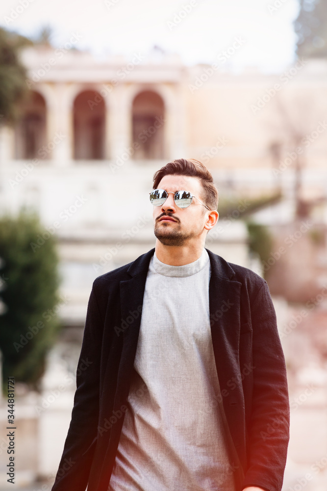 Stylish man walking with sunglasses and modern hairstyle and beard, looking up. Outdoor in the street. White t-shirt and black jacket. Stylish man walking with sunglasses and modern hairstyle and beard, looking up. Outdoor in the street. White t-shirt and black jacket.