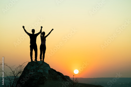 Man and woman hikers standing on a big stone at sunset in mountains. Couple raising up hands on high rock in evening nature. Tourism, traveling and healthy lifestyle concept.